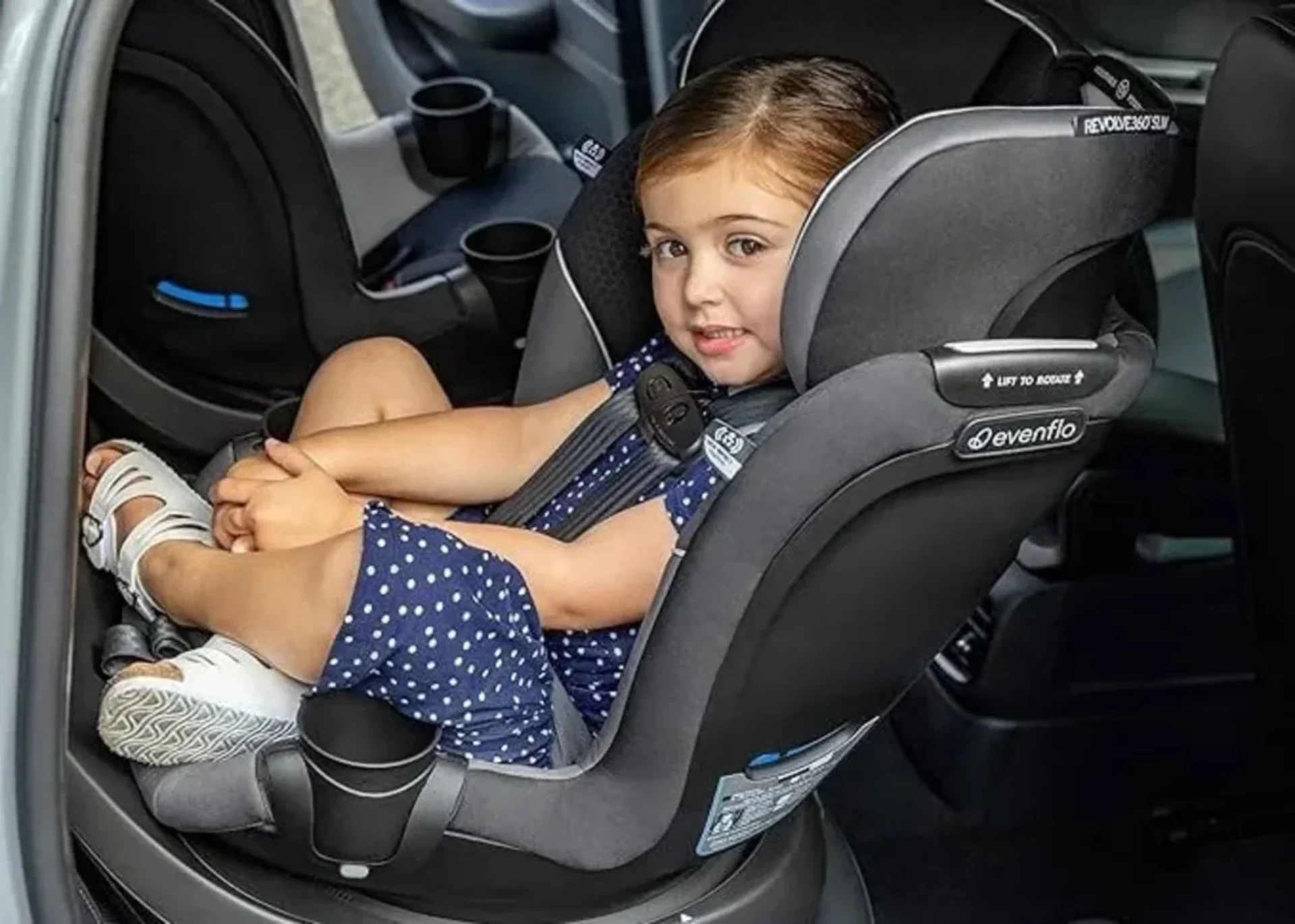 Smiling toddler girl in blue polka dot dress seated safely in a dark gray Evenflo Revolve360 Slim convertible car seat inside