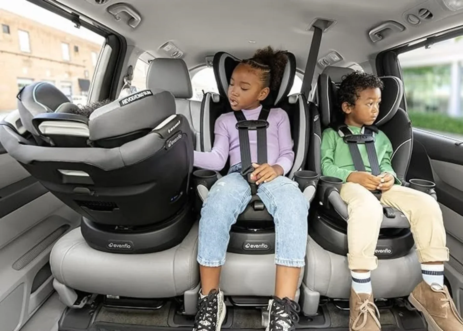 Two children secured in Evenflo Revolve Slim convertible car seats in a minivan back row with an infant seat on the left