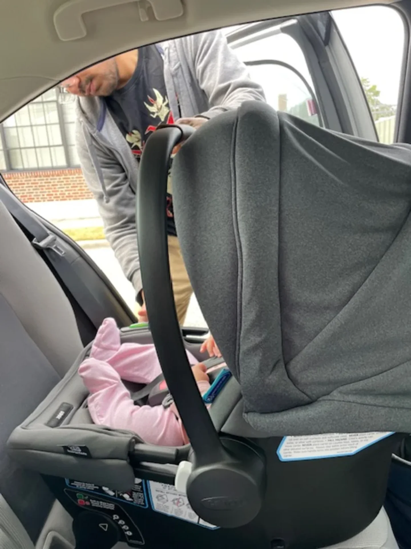 Adult securing a baby in a gray Graco GoMax baseless infant car seat inside a vehicle backseat, baby wearing pink outfit