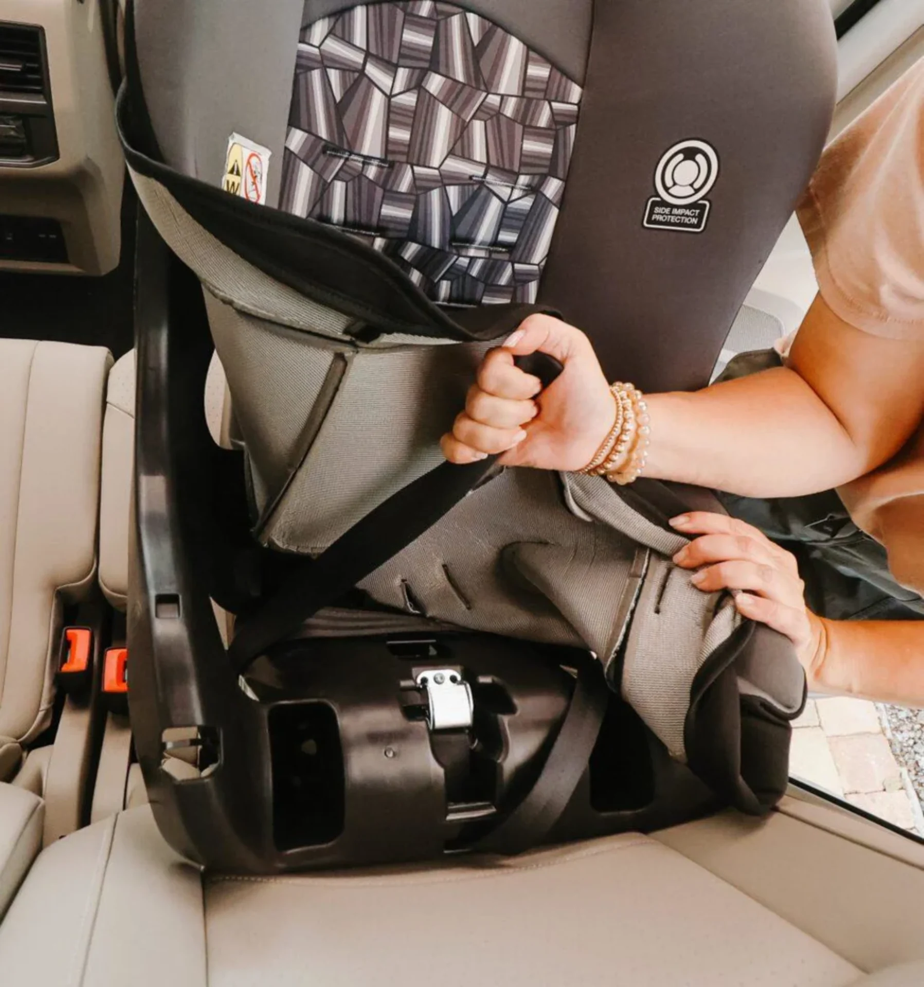 Woman installing a Cosco Scenera Next car seat with side impact protection in a beige vehicle rear seat, adjusting harness st