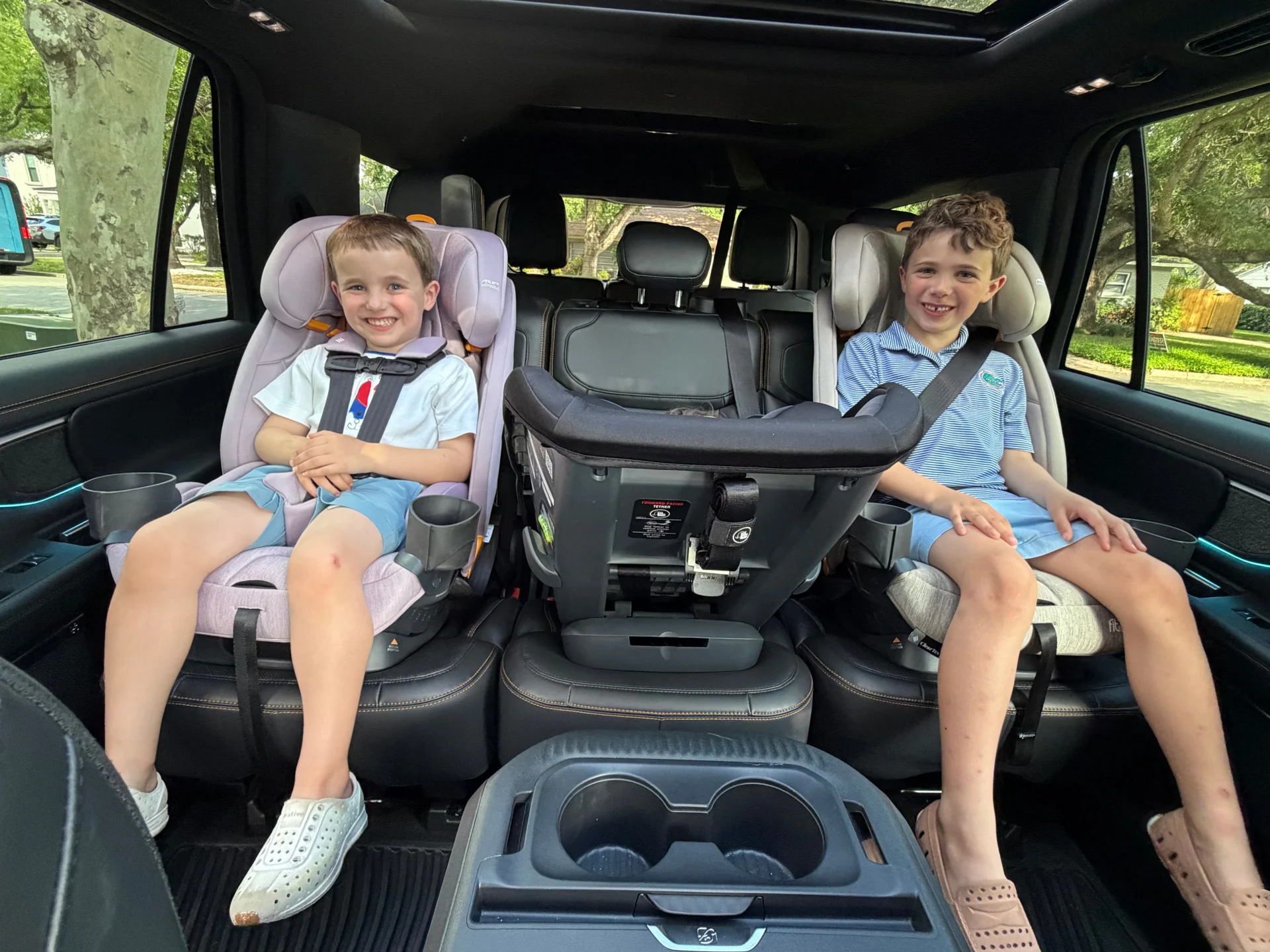 Two smiling young boys securely buckled into Chicco Fit3x convertible car seats in the spacious second row of a modern SUV