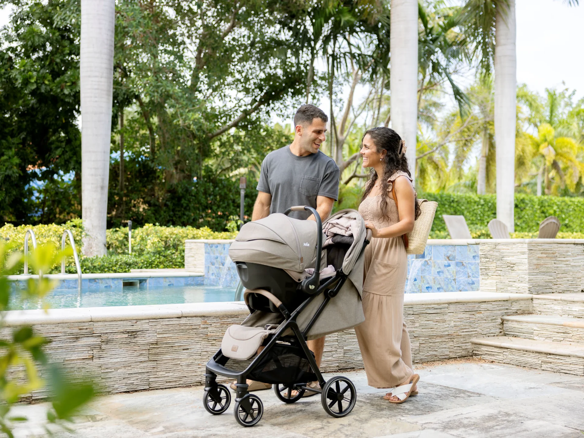 Smiling couple strolling with a Joie travel system stroller along a sunlit tropical resort poolside surrounded by palm trees