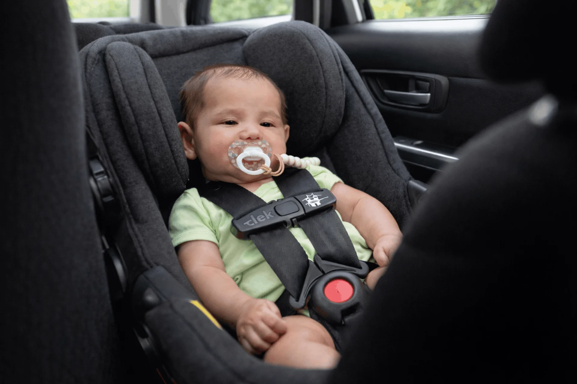 Smiling infant with pacifier wearing green onesie safely buckled into a gray Clek rear-facing car seat in a vehicle backseat