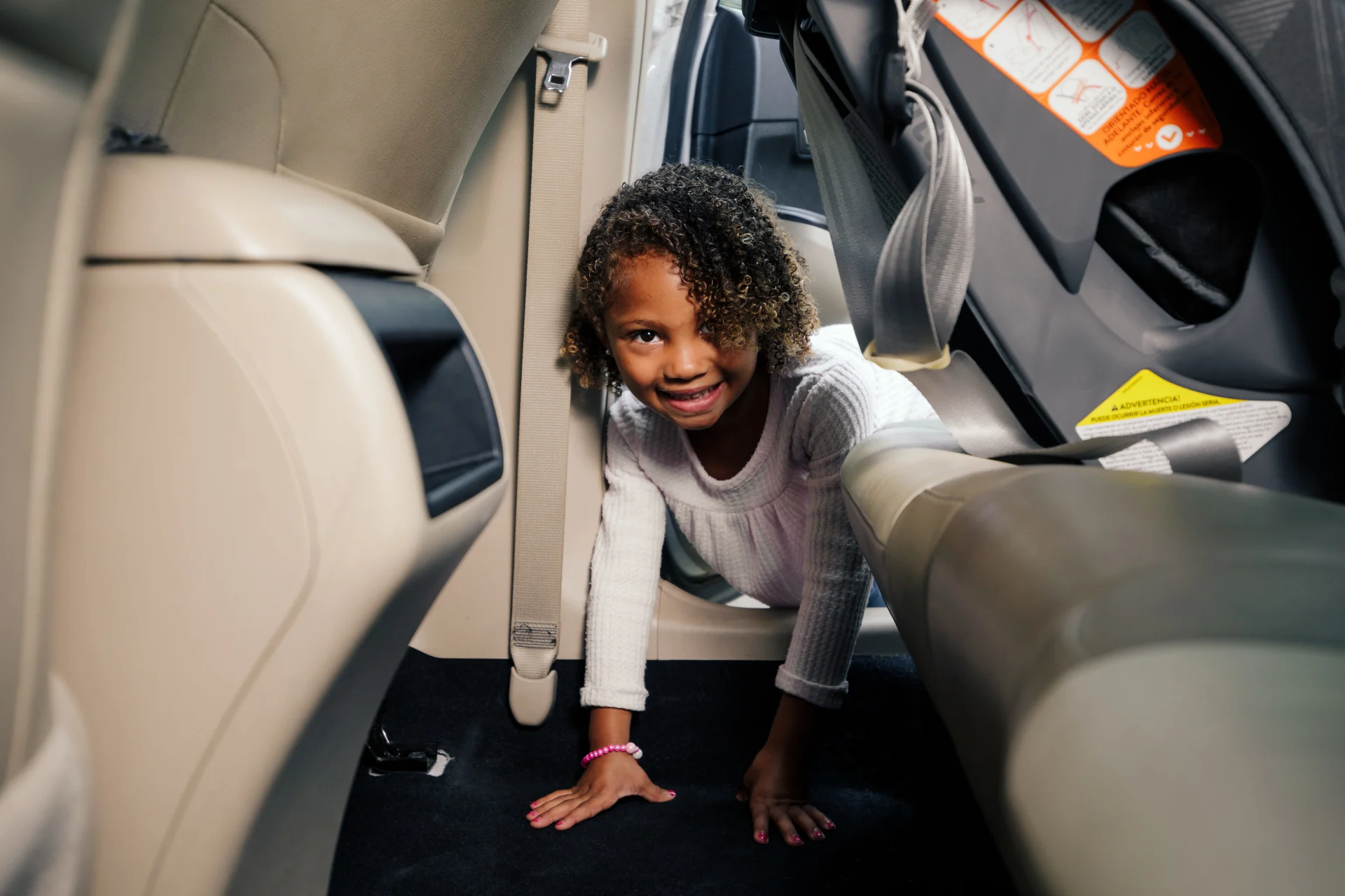 Smiling young girl with curly hair crawling on the floor of a car backseat between beige leather seats near a child car seat