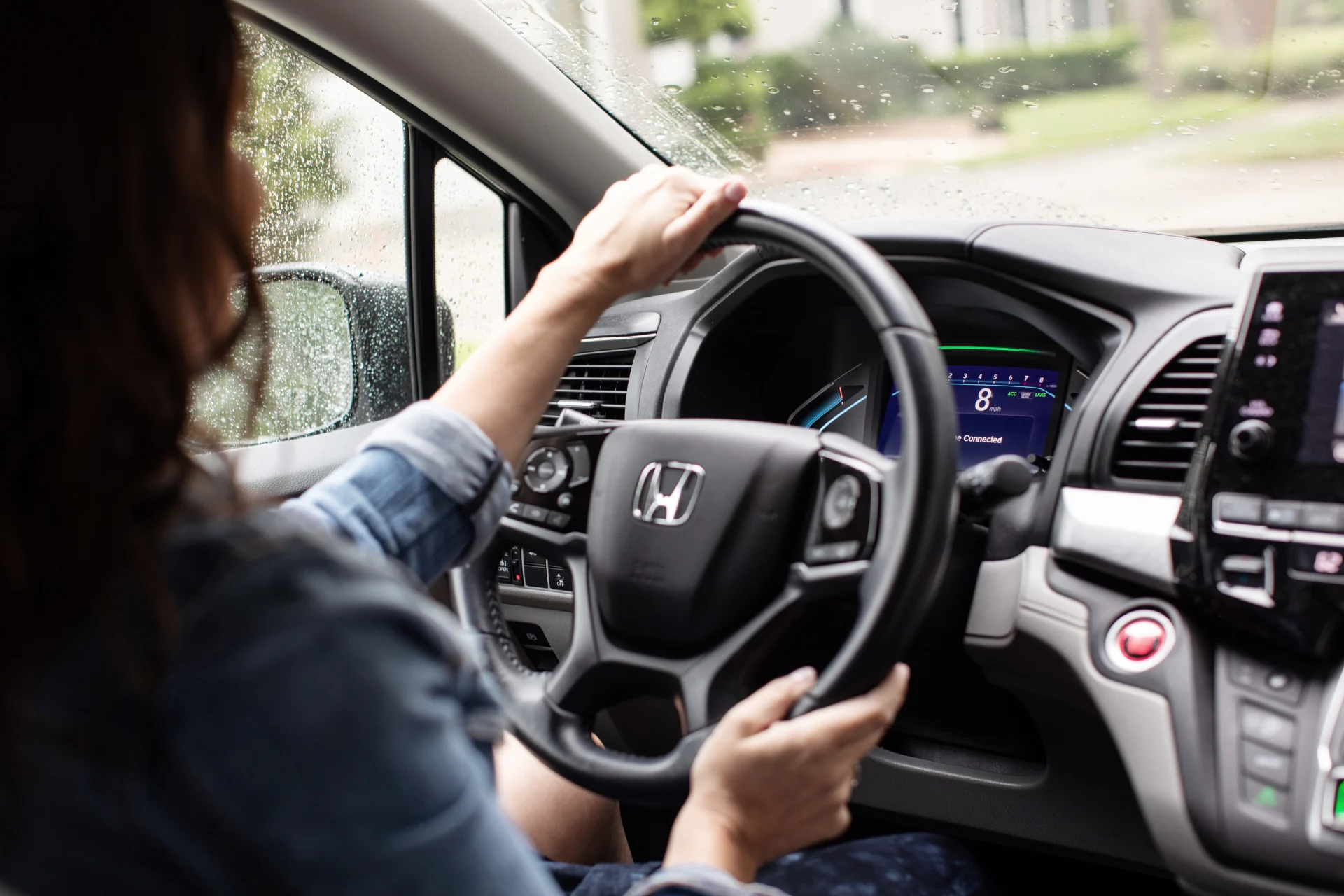 Woman in denim jacket gripping Honda steering wheel while driving in rainy weather, digital dashboard and rain-spotted window