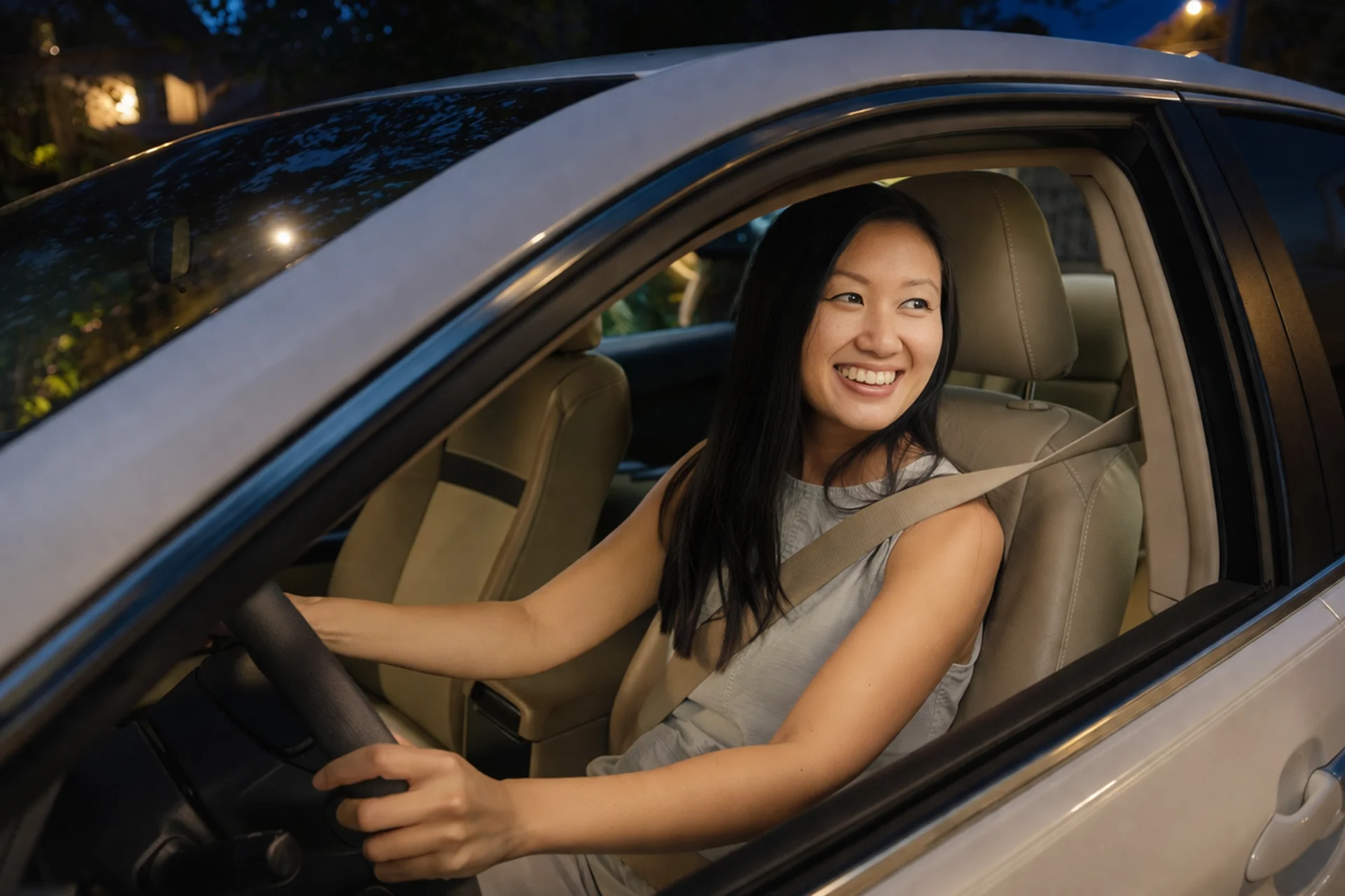 Smiling young woman with long dark hair sitting in driver's seat of silver car at night, wearing seatbelt, beige leather inte