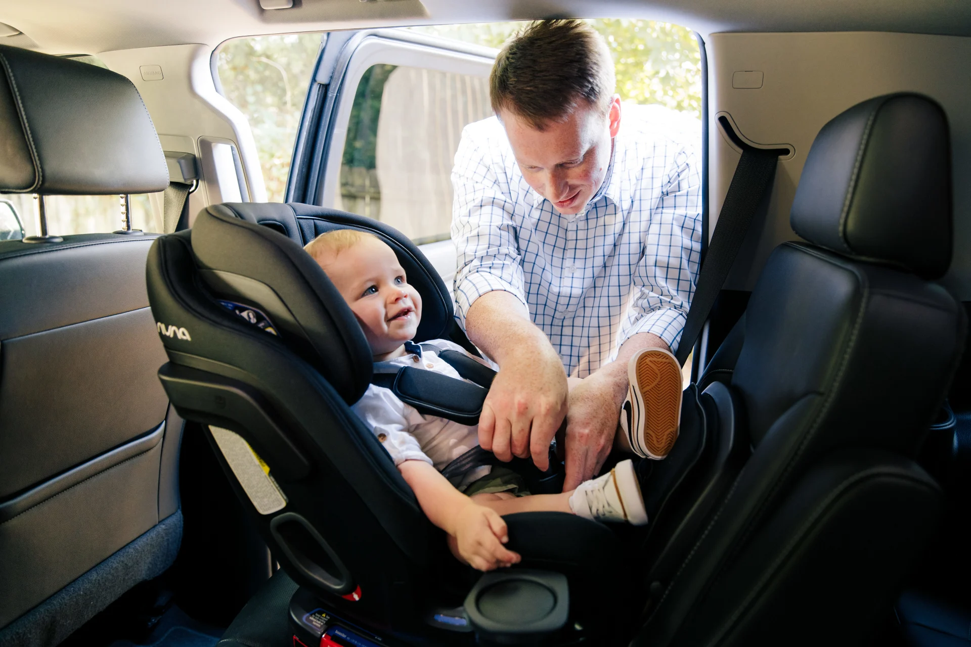 Father in plaid shirt buckling a smiling toddler into a black Nuna convertible car seat in the rear of an SUV