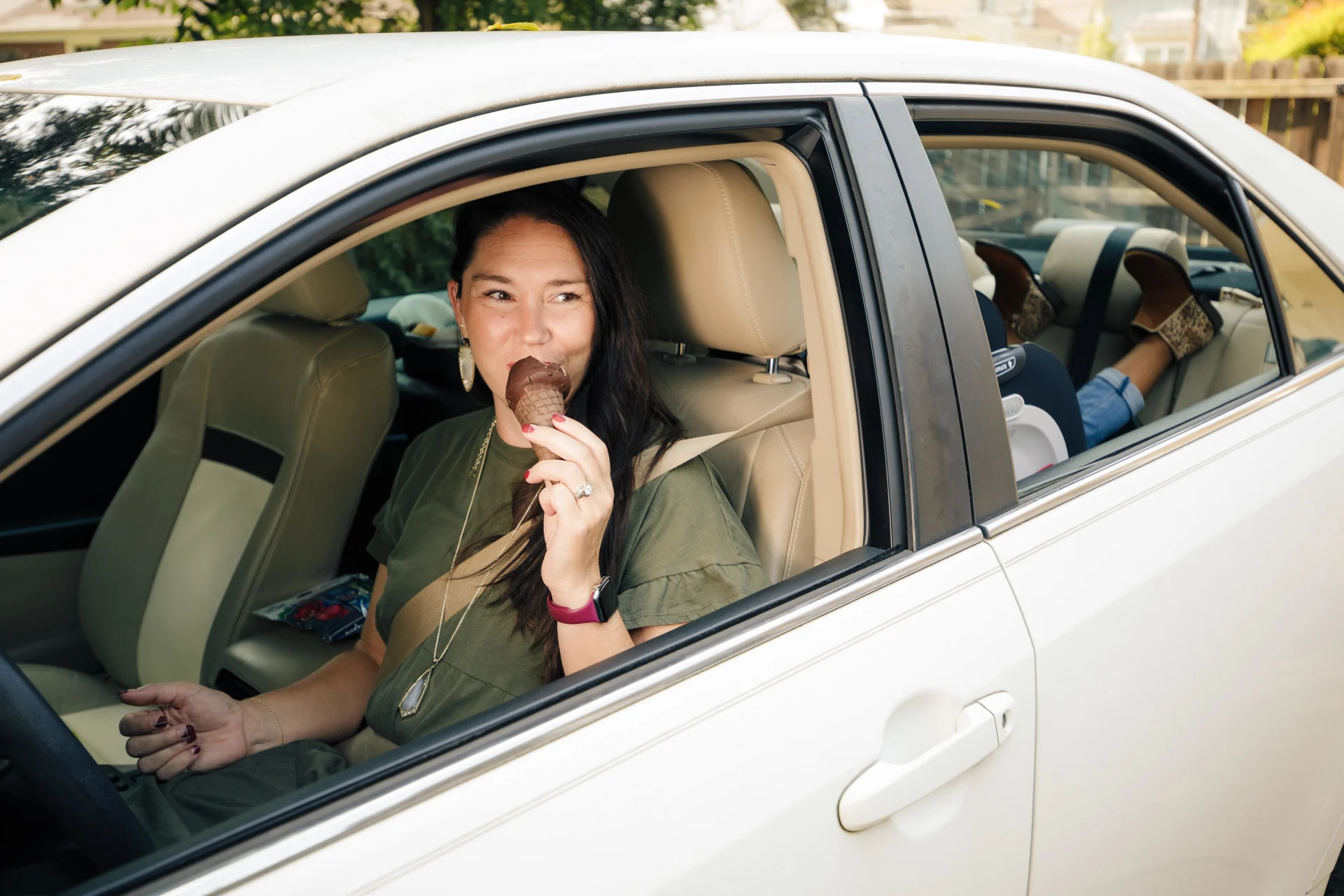Smiling dark-haired woman eating a chocolate-dipped ice cream cone in the driver seat of a white car with the window down