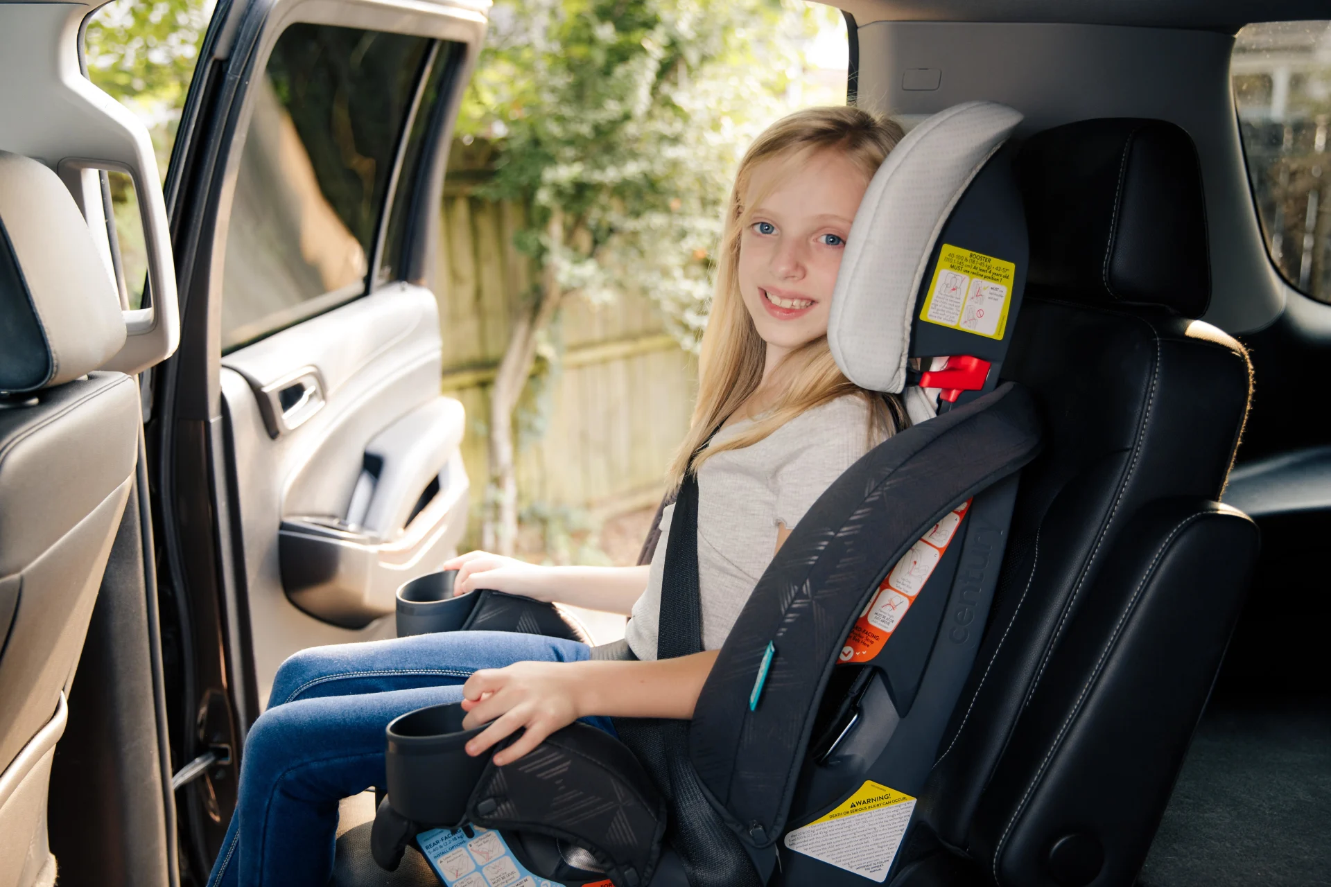 Smiling blonde girl with blue eyes seated securely in a black booster car seat with safety harness in a vehicle backseat