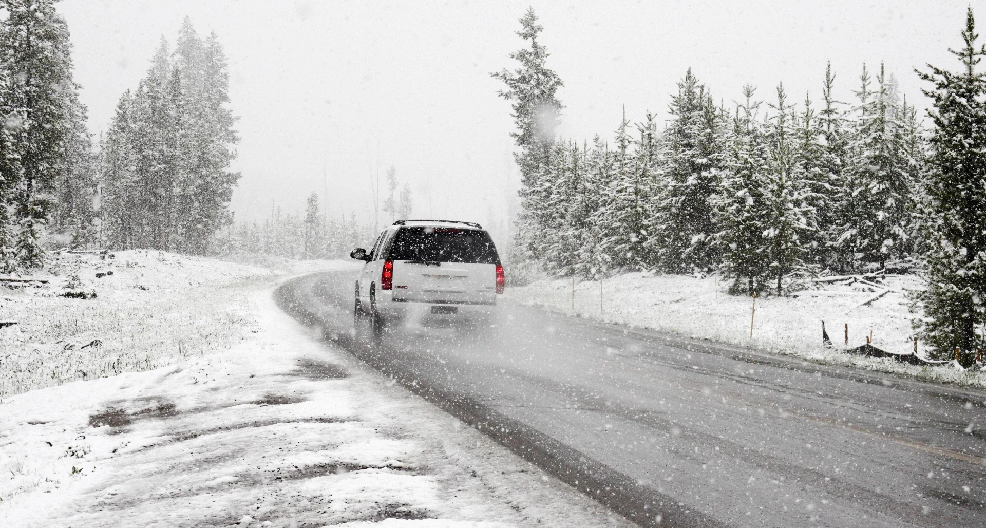 White GMC SUV navigating a slippery snow-covered rural road through snow-dusted pine forest during heavy snowfall with low vi