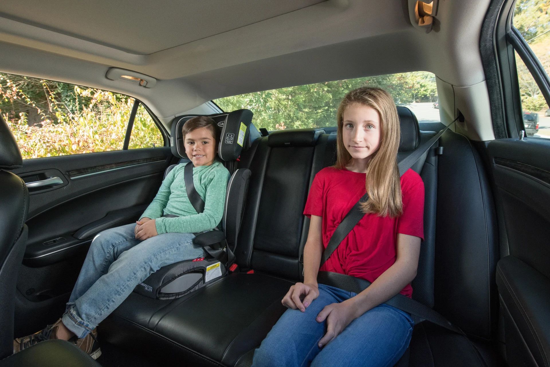 Two children safely buckled in a car backseat, younger child in a black booster seat wearing green shirt, older girl in red s