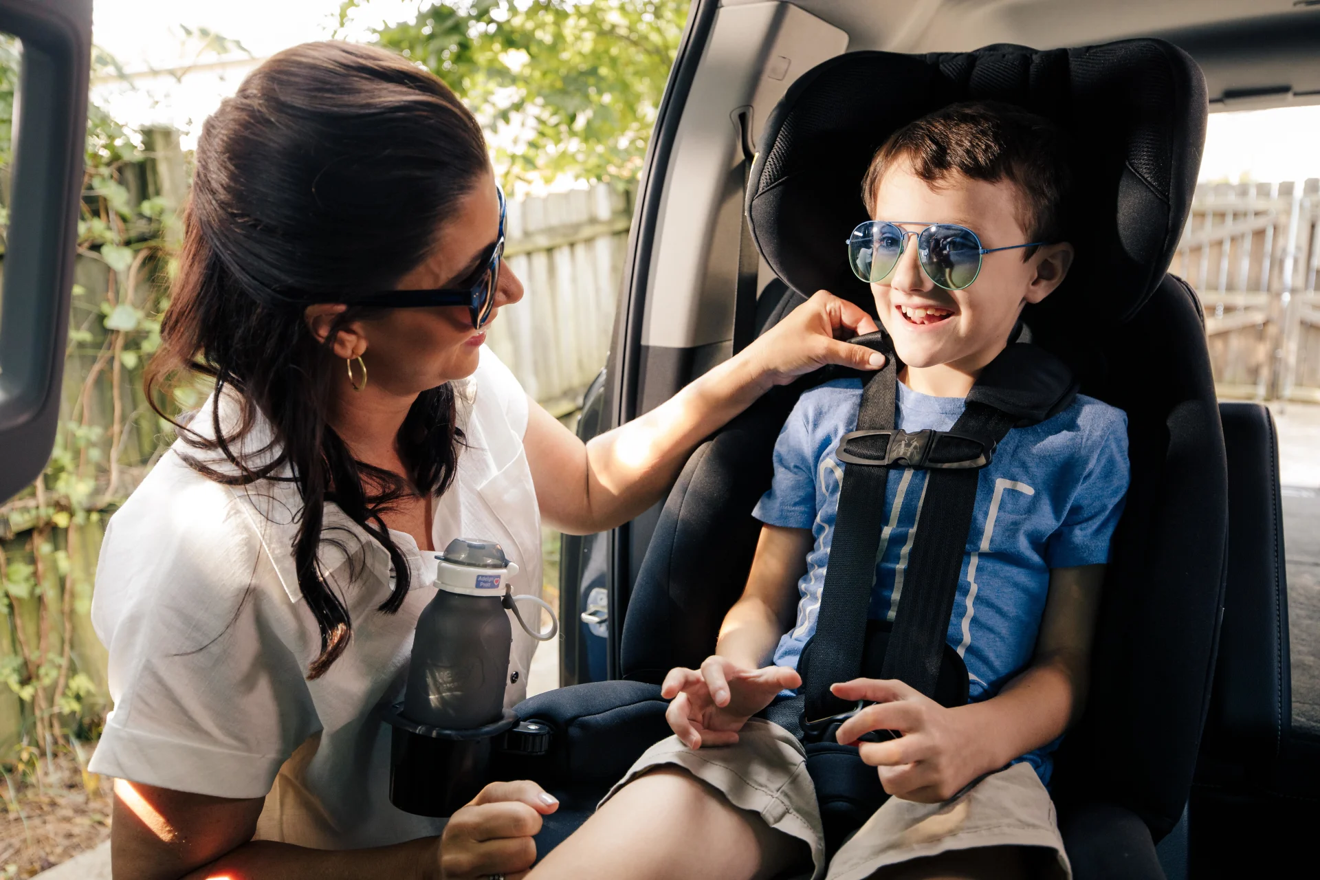 A smiling mother in sunglasses buckles her happy young boy wearing aviator sunglasses into a black car seat on a sunny day