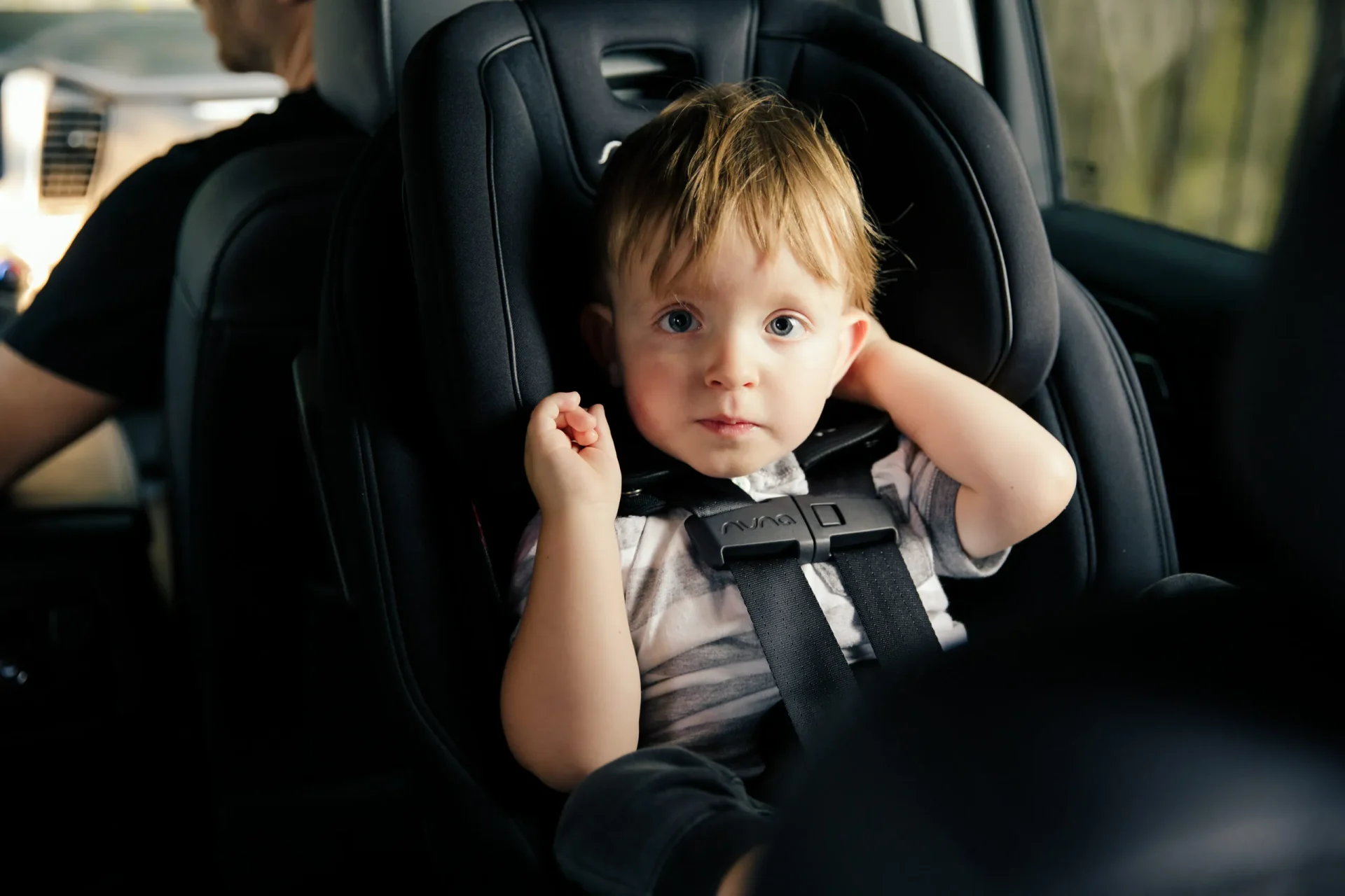 Young toddler with blonde hair and blue eyes safely buckled into a black Nuna car seat in a vehicle backseat, looking at came