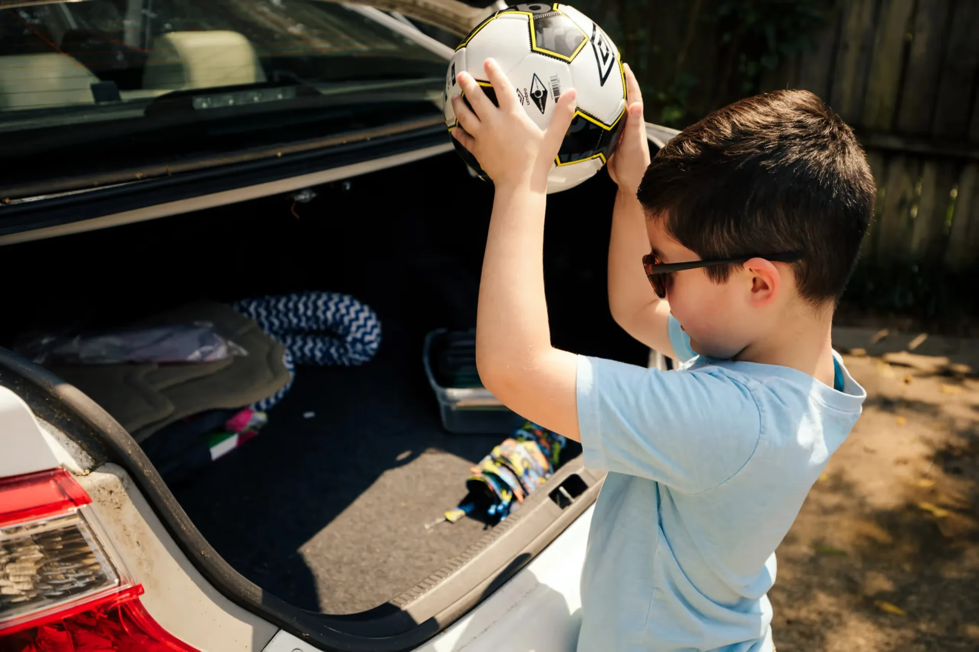 Young boy wearing sunglasses and a light blue shirt lifting a soccer ball into an open white car trunk while packing for a ro