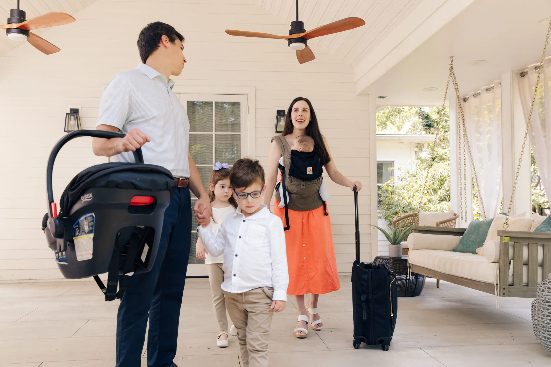 Young family with three children departing a white shiplap porch, father carrying Britax infant car seat, mother wearing baby
