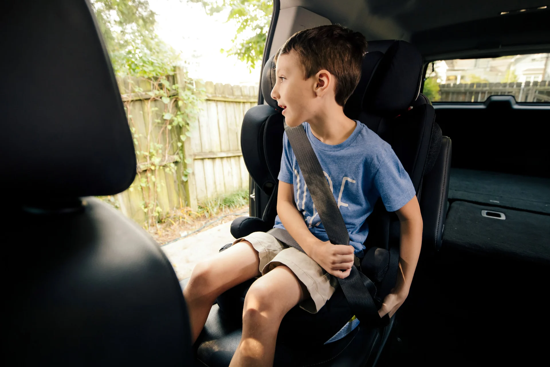Young boy in blue t-shirt secured with seatbelt in a black high-back booster seat, gazing out the car window
