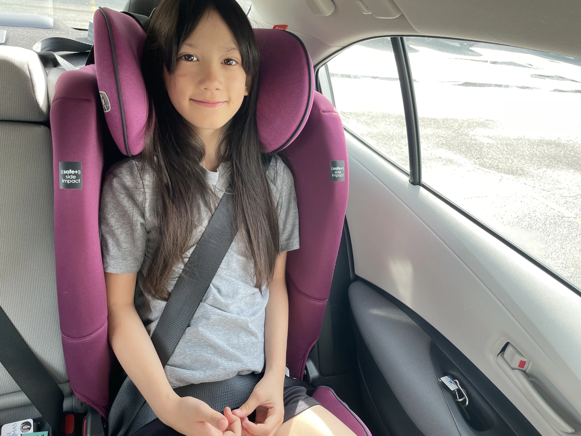 Young girl with long dark hair sitting safely buckled in a purple side-impact booster seat in a car backseat, smiling at came