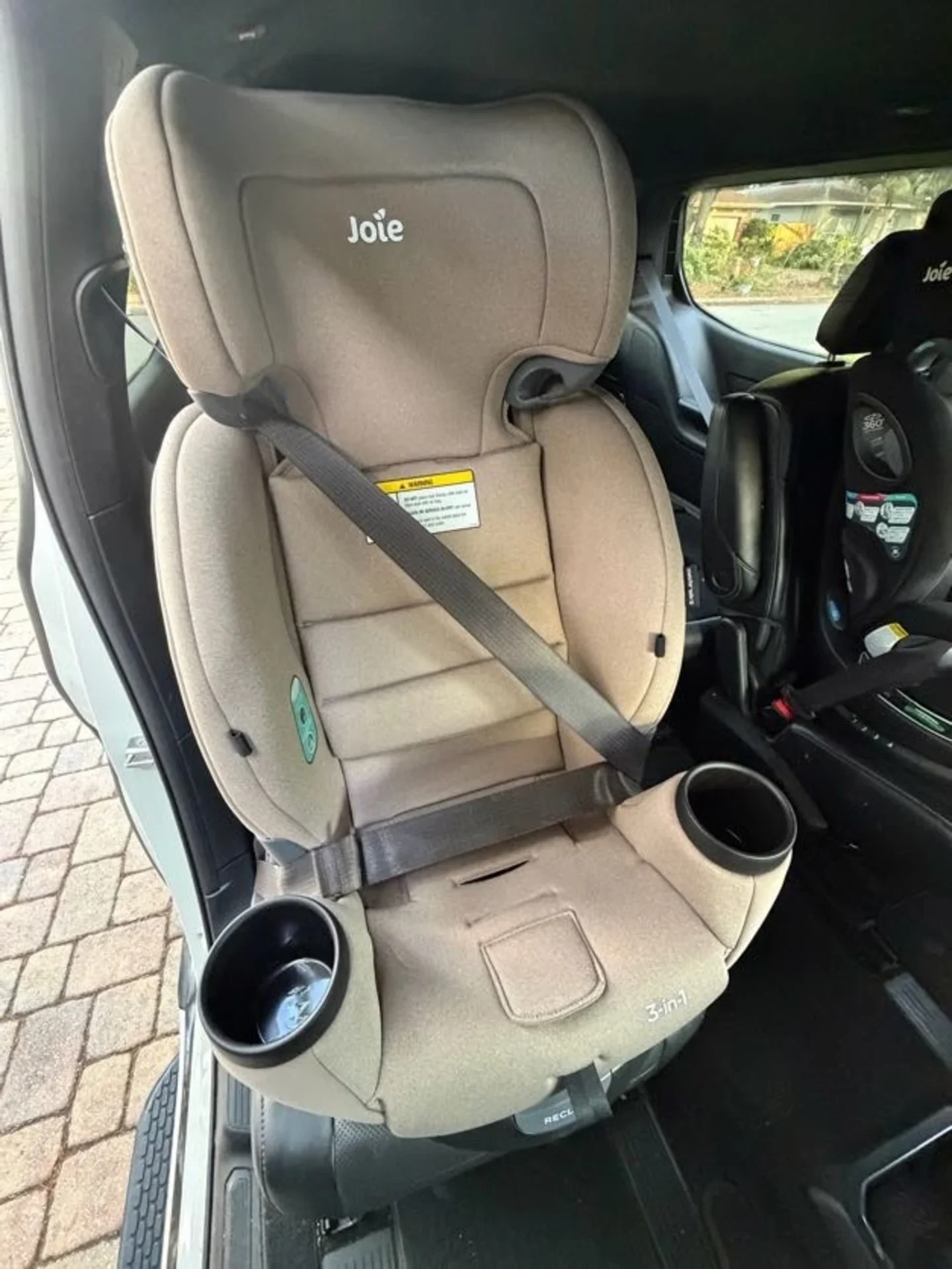 Joie 3-in-1 Child Car Seat Installed in Vehicle Joie 3-in-1 beige child car seat with dual cup holders and grey seatbelt routing installed in rear of a vehicle next to a Joi