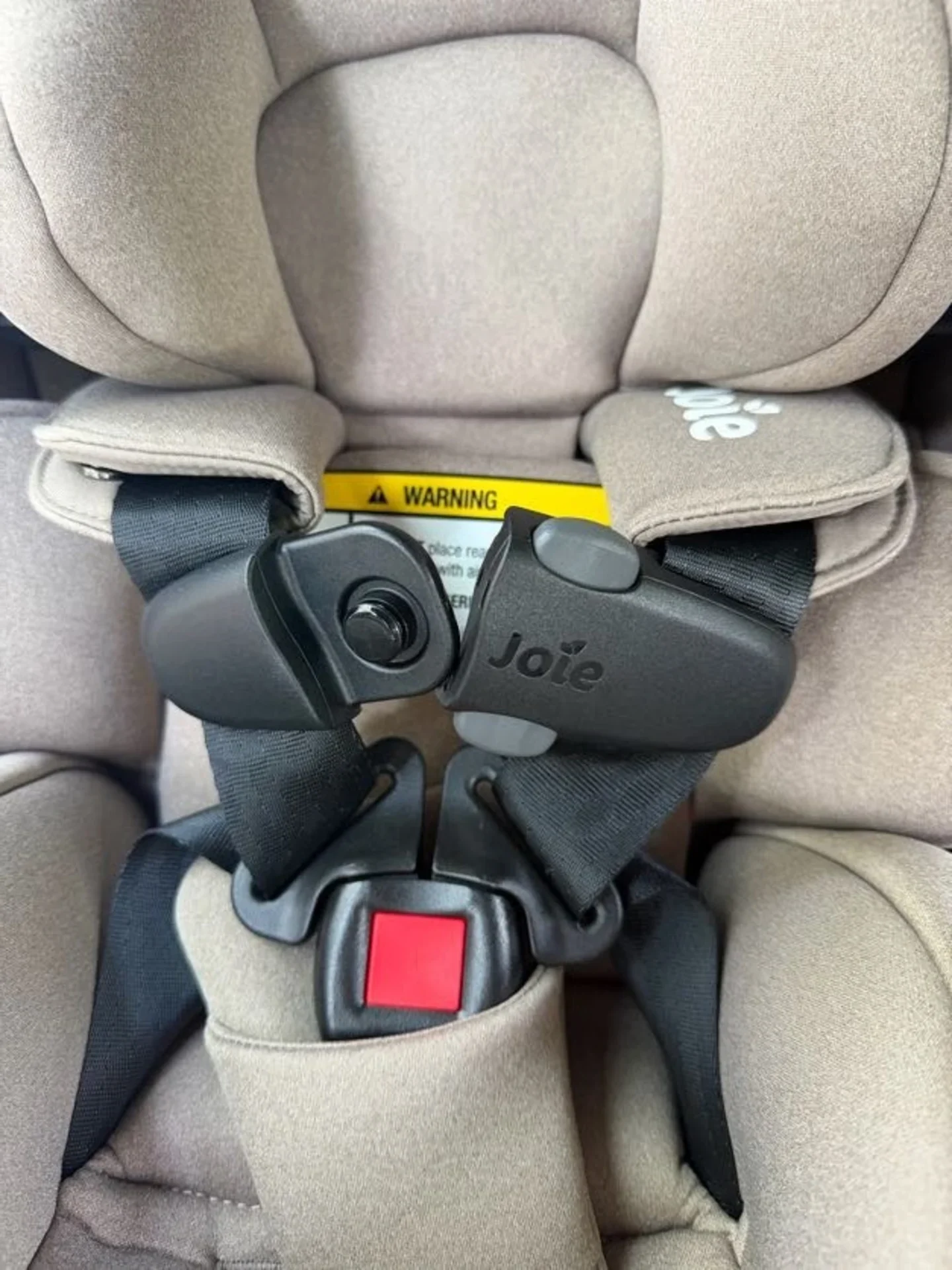 Joie Spin SI Harness and Buckle Detail Close-up of Joie Spin SI car seat 5-point harness with black chest clip, red release button, and yellow warning label on beig