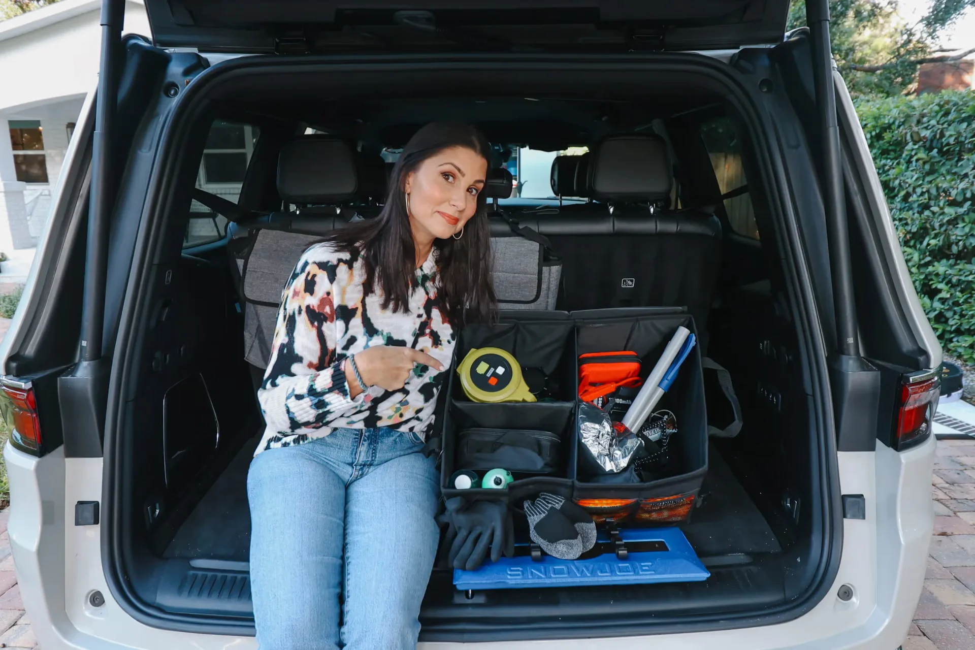 Woman sitting in open SUV trunk pointing to organized storage organizer filled with tape measure, gloves, emergency supplies,