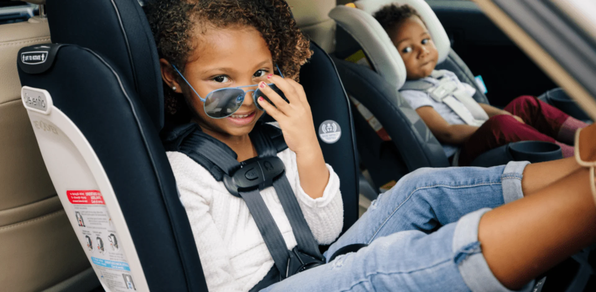 Young girl wearing blue aviator sunglasses smiling in a navy Evenflo car seat with a toddler secured in a rear-facing seat be