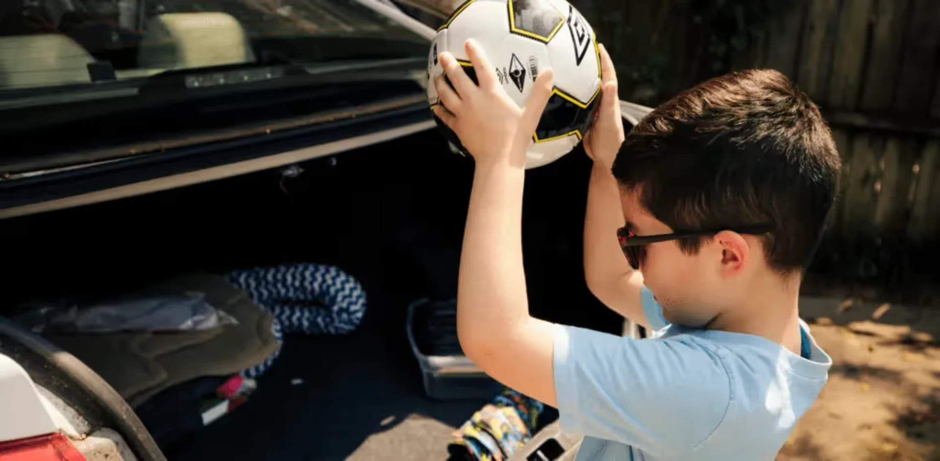 Young boy wearing sunglasses and a light blue shirt lifting a black and white soccer ball above his head near an open car tru