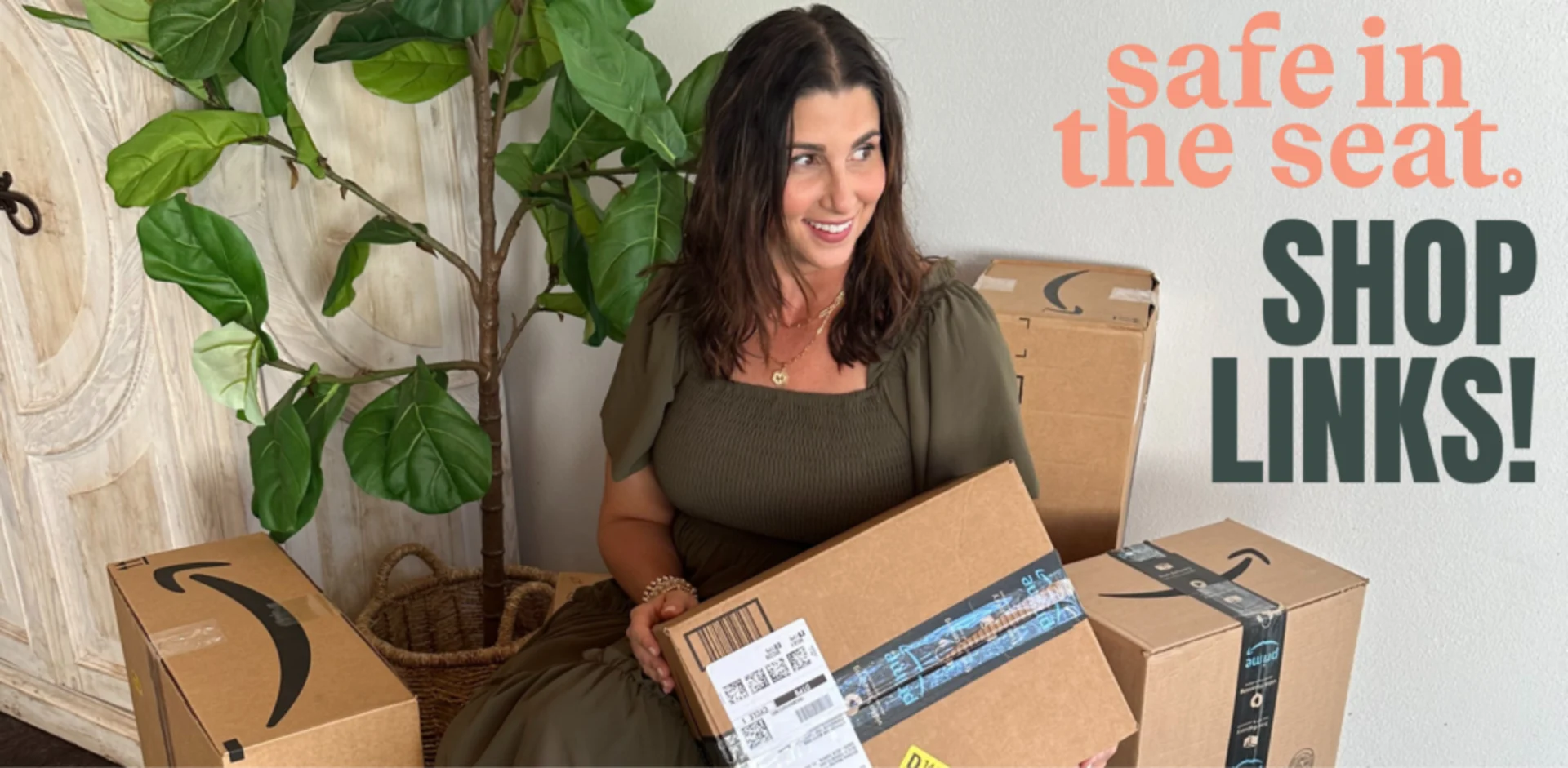 Smiling brunette woman in olive dress holding Amazon Prime packages surrounded by delivery boxes next to a fiddle leaf fig tr