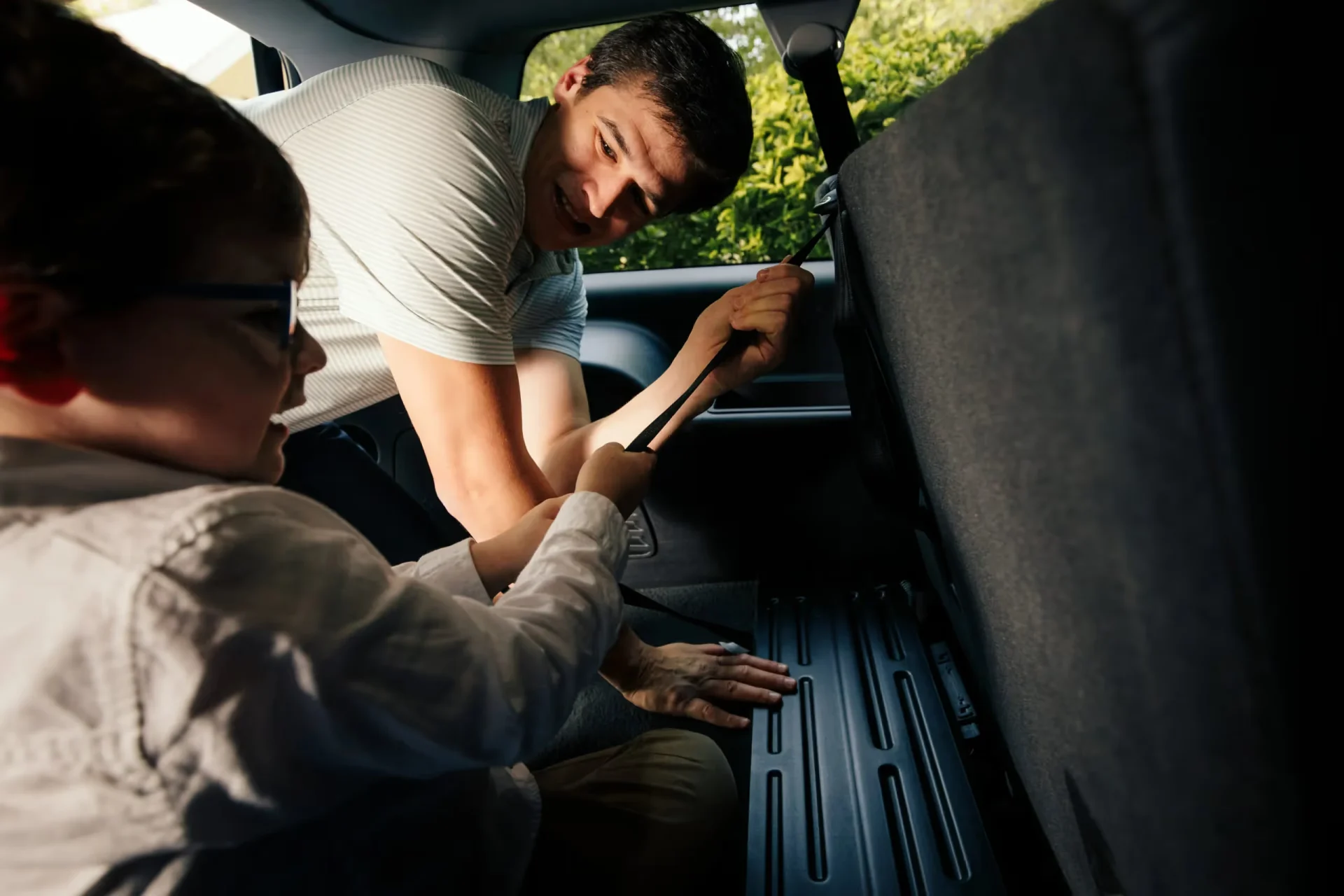 Smiling father leaning into car backseat helping child secure a top tether strap on a child safety seat