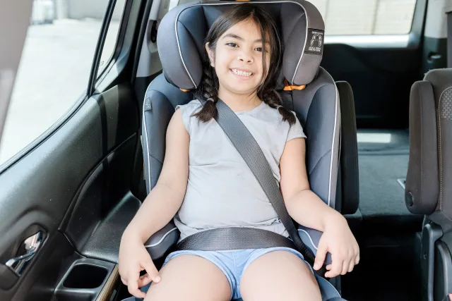 Smiling young girl with braids sitting securely in a gray high-back booster car seat with seatbelt fastened inside a vehicle