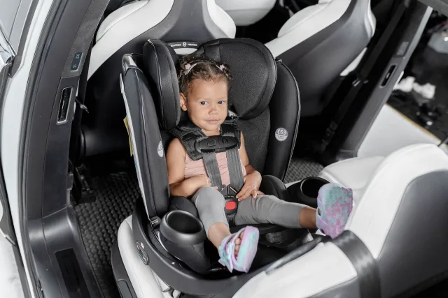 Young toddler girl with braided hair and pink hair clips secured in a black swivel convertible car seat with 5-point harness