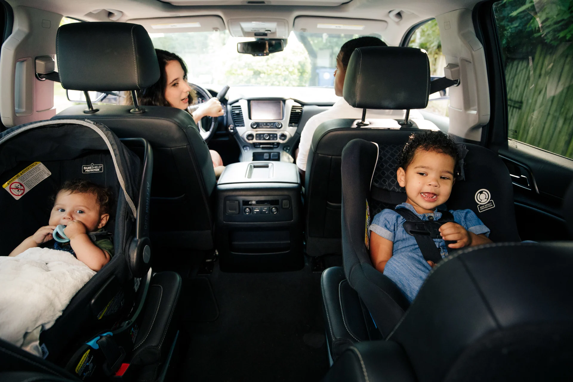 Two young children secured in black car seats in the backseat of an SUV while two adults sit in the front seats during a fami