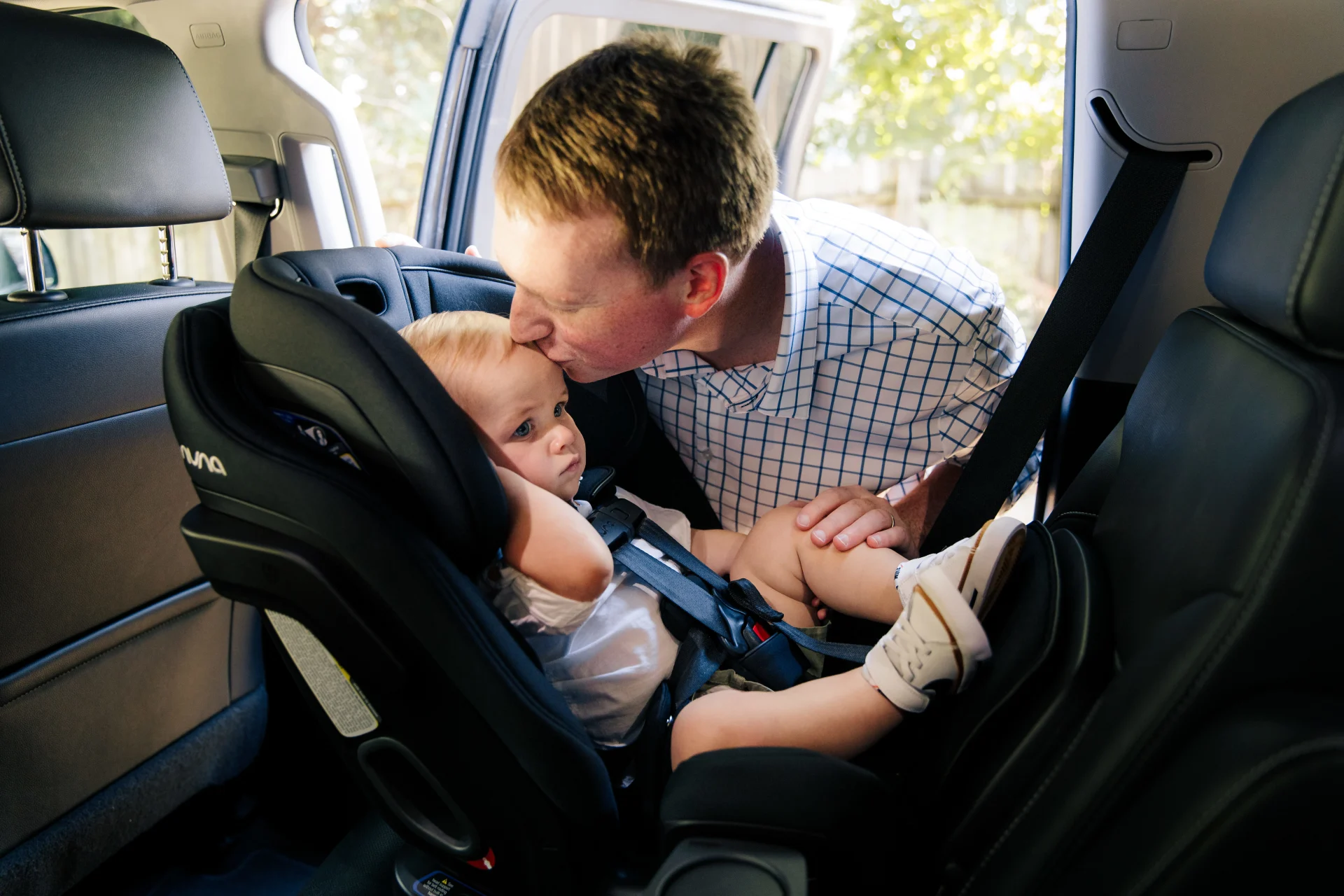 Father in plaid shirt leaning into car backseat to kiss blonde baby securely buckled in a Nuna rear-facing car seat