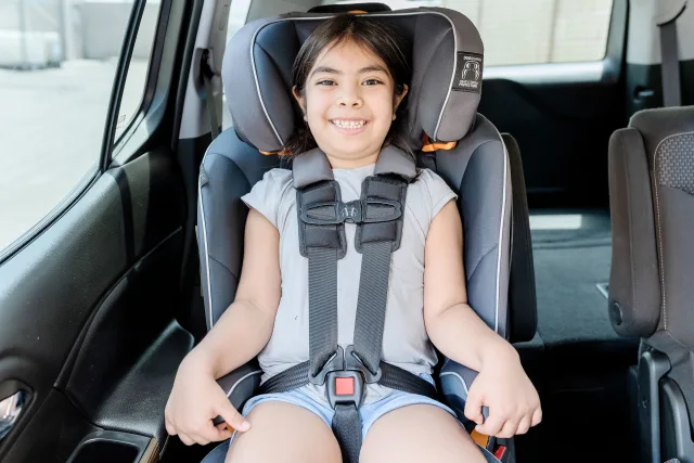 Smiling young girl safely buckled into a gray forward-facing car seat with a 5-point harness inside a vehicle