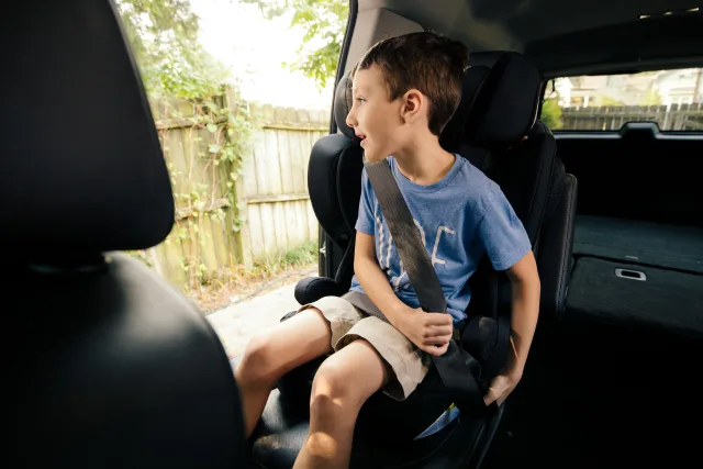 Young boy in blue t-shirt seated securely in black booster car seat with seatbelt fastened, looking out car window