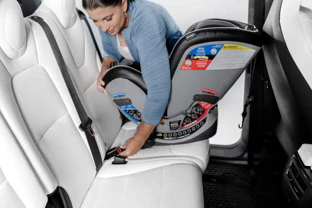 Woman in blue cardigan securing a Chicco convertible car seat with the LATCH system into a white leather vehicle rear seat