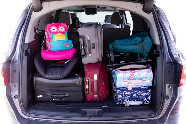 Fully packed SUV trunk with multiple suitcases, a pink child car seat topped with a colorful owl backpack, folded stroller, a