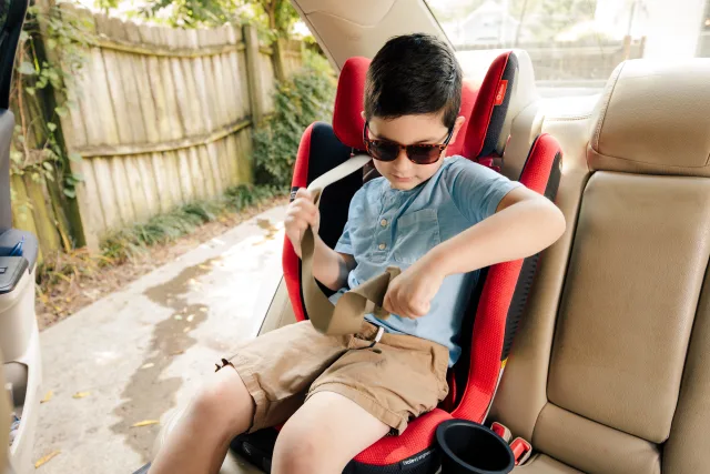 Young boy in sunglasses and blue shirt independently buckling a seatbelt in a red booster car seat in a vehicle backseat