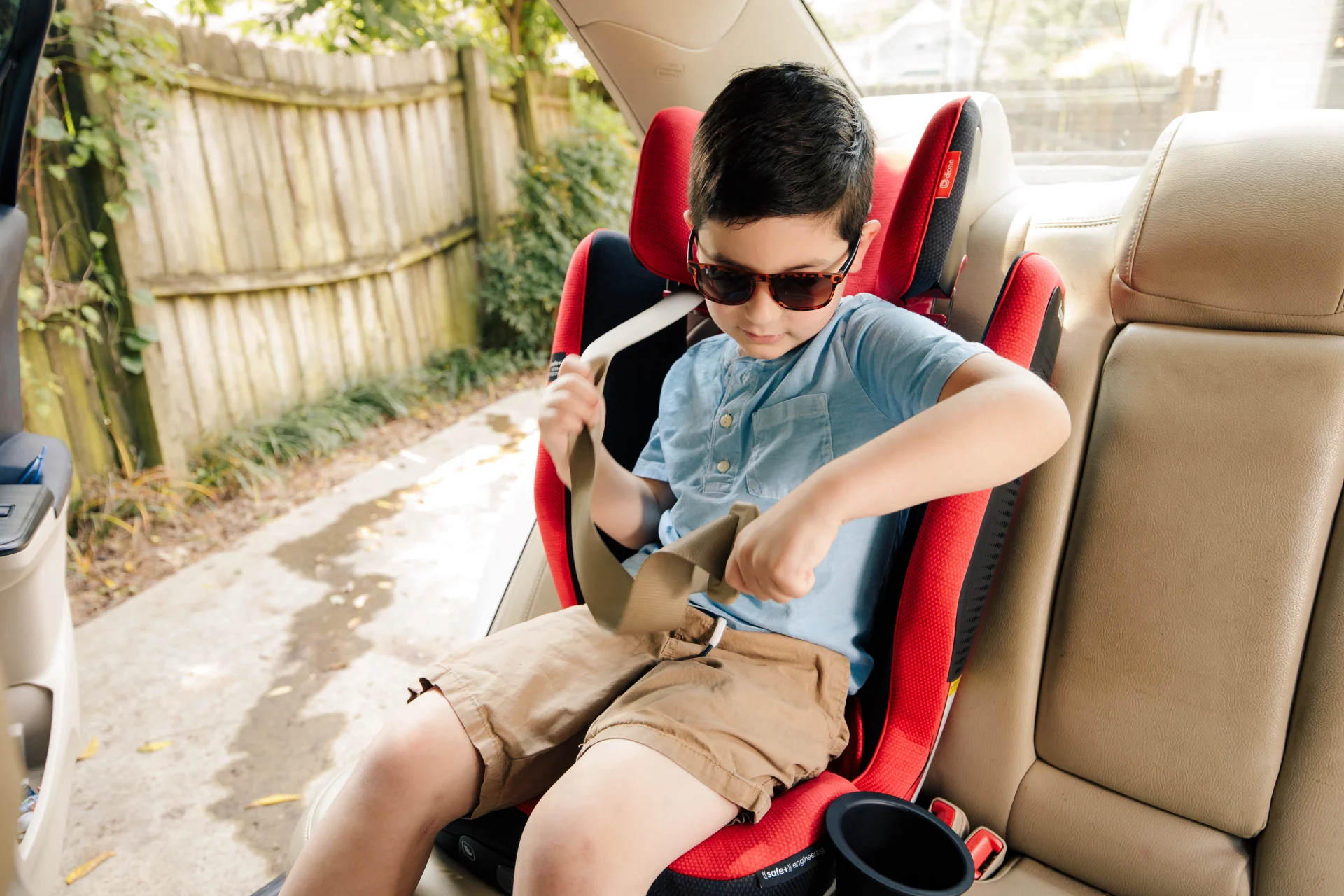 Young boy in sunglasses and blue shirt independently buckling a seatbelt in a red booster car seat in a vehicle backseat