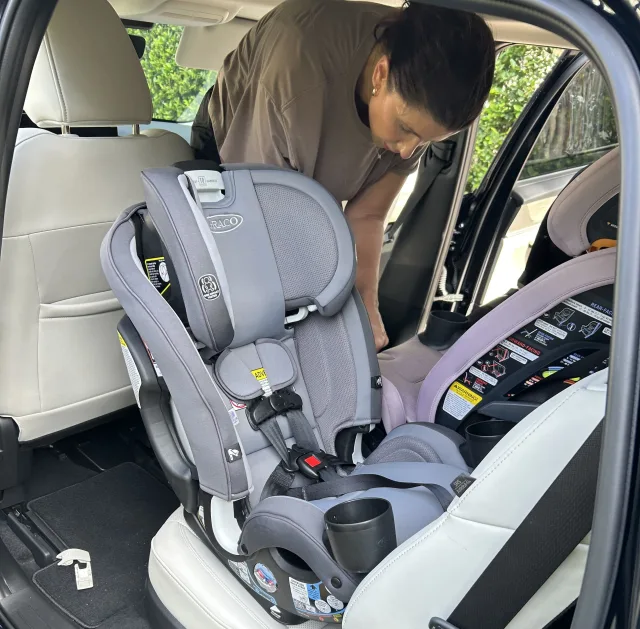 Woman leaning into a car backseat installing a gray Graco convertible car seat next to a purple rear-facing infant car seat