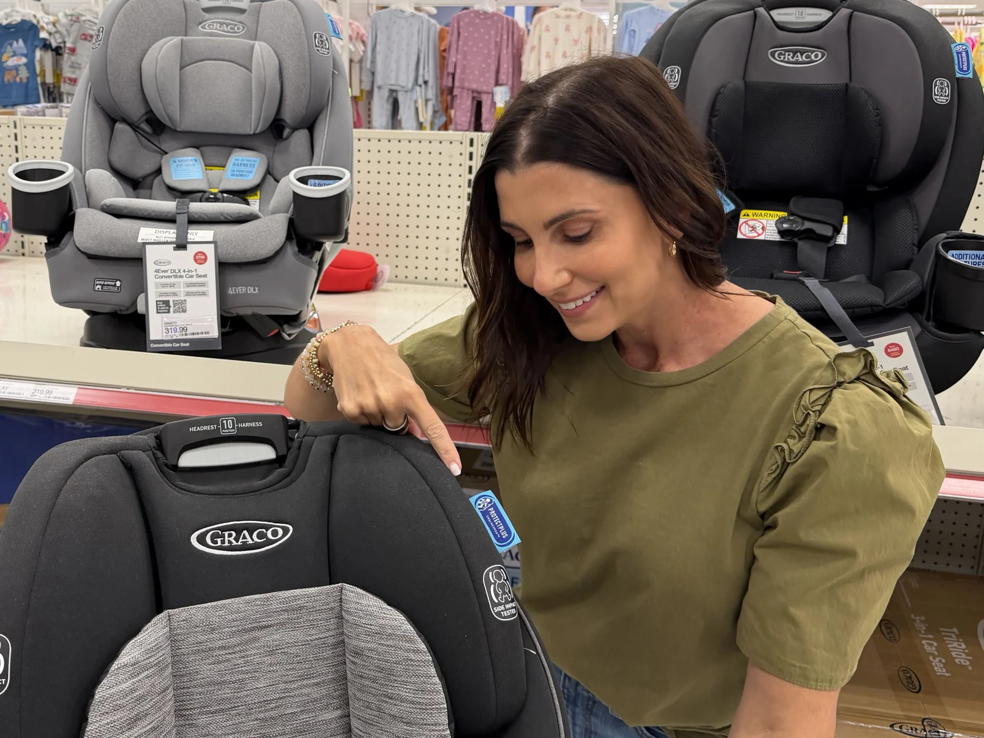Smiling brunette woman in olive green top pointing at Graco convertible car seat headrest adjuster in Target baby aisle