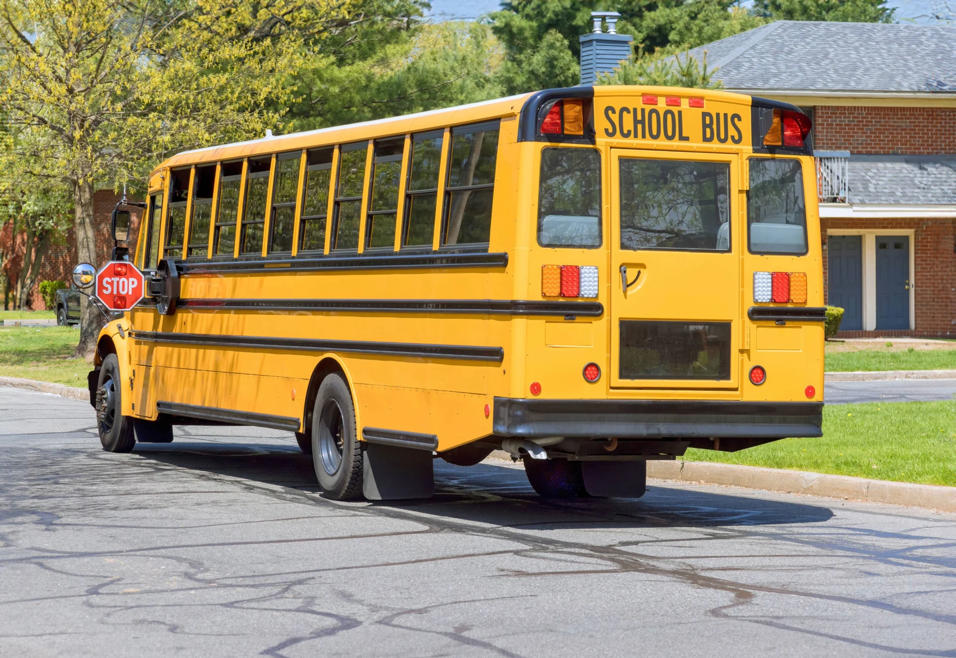 Yellow school bus stopped on a residential street with red stop sign arm extended, brick houses and green trees visible in ba