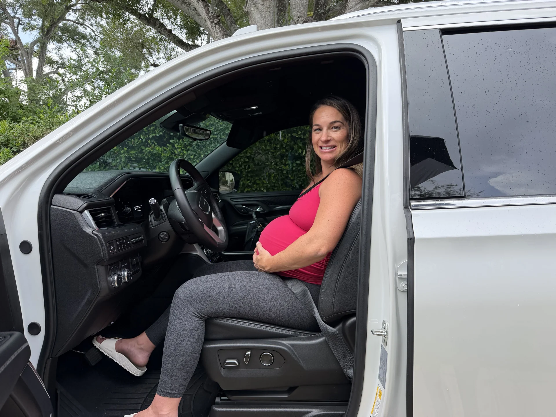 Smiling pregnant woman in pink tank top and gray leggings sitting in open driver seat of white truck, cradling her baby bump
