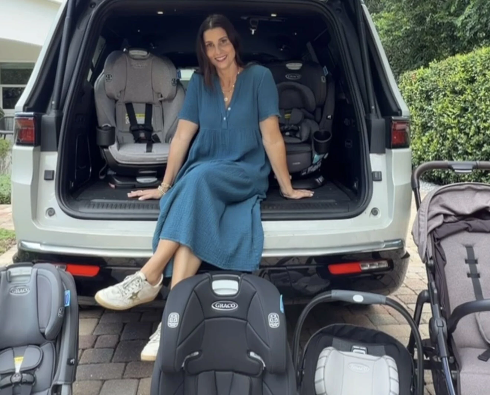 Smiling woman in blue dress sitting in open SUV trunk surrounded by multiple Graco car seats and a stroller for comparison