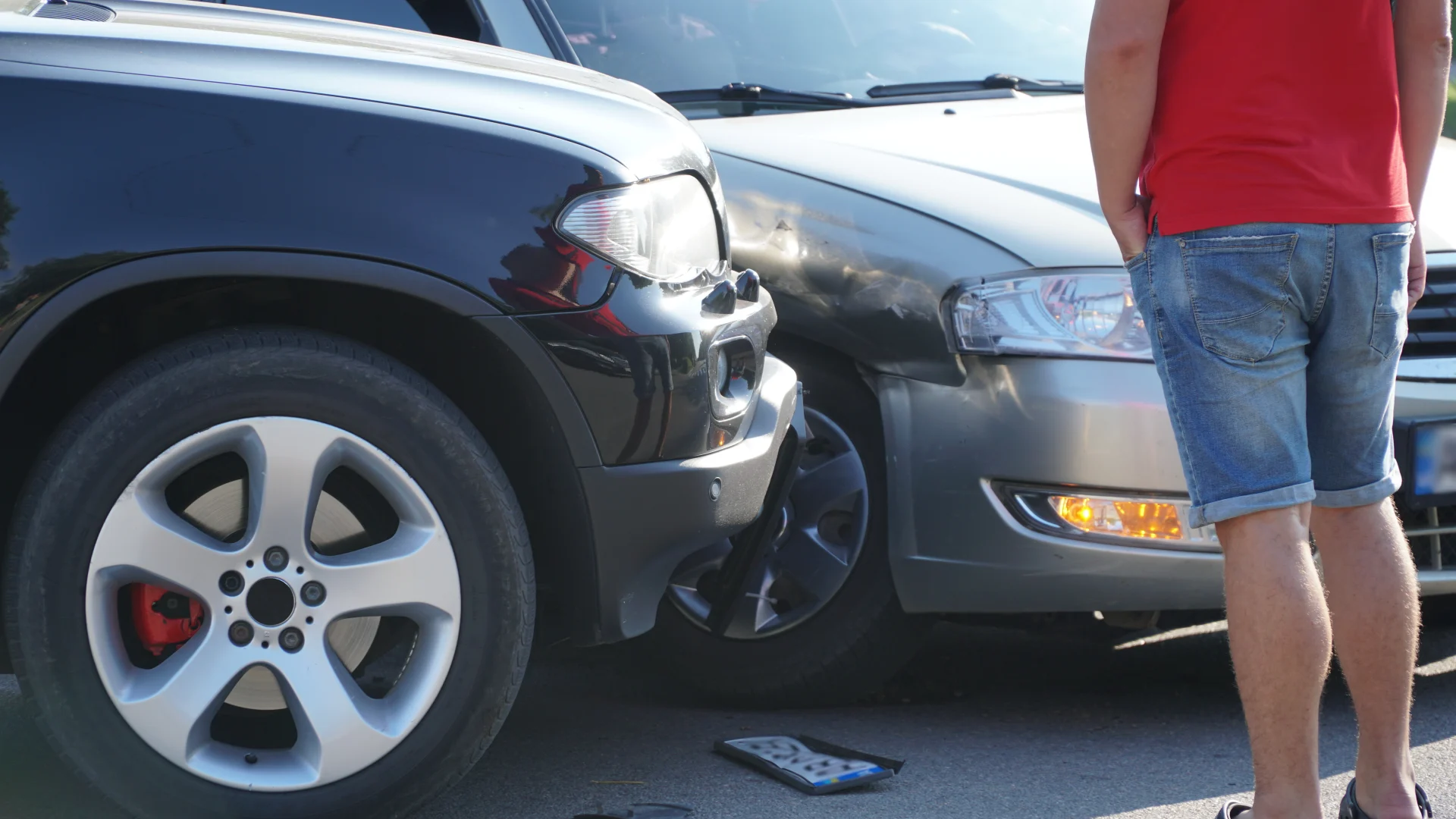 Person in red shirt and denim shorts standing beside two cars with crumpled front-end collision damage on a sunny day, phone