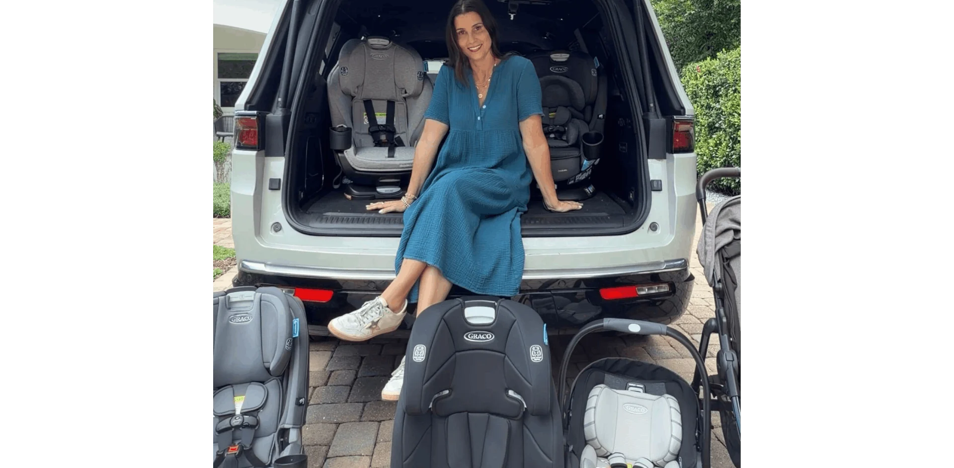 Smiling woman in teal dress sitting in open white SUV trunk surrounded by multiple Graco car seats and infant carriers outdoo