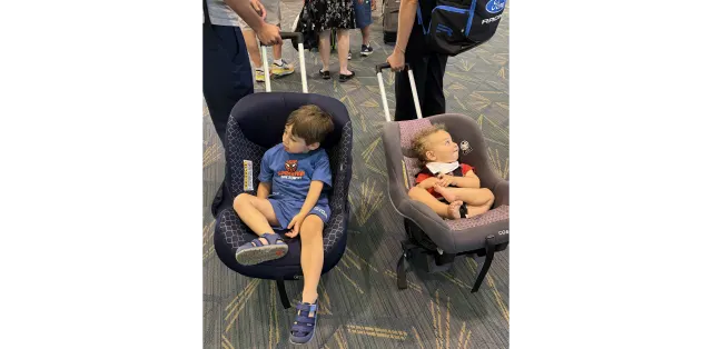 Toddler in blue Spider-Man shirt and infant in red outfit seated in separate car seats on patterned airport terminal floor su