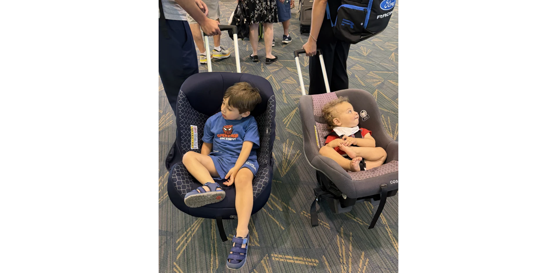 Toddler in blue Spider-Man shirt and infant in red outfit seated in separate car seats on patterned airport terminal floor su