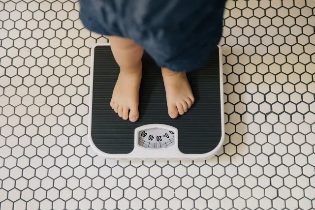 Overhead view of a young child in blue pants standing barefoot on a white and black analog bathroom scale on hexagon tile flo