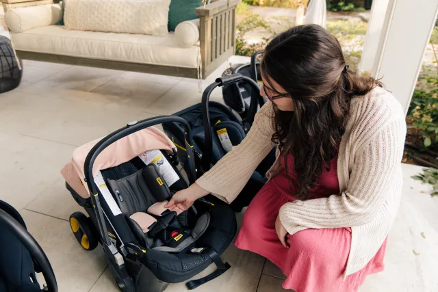 Pregnant woman in red dress and cream cardigan crouching to inspect a pink and black Doona infant car seat stroller on a porc