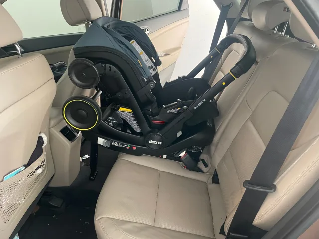 Doona X infant car seat and stroller combo installed rear-facing on the middle seat of a beige leather SUV interior